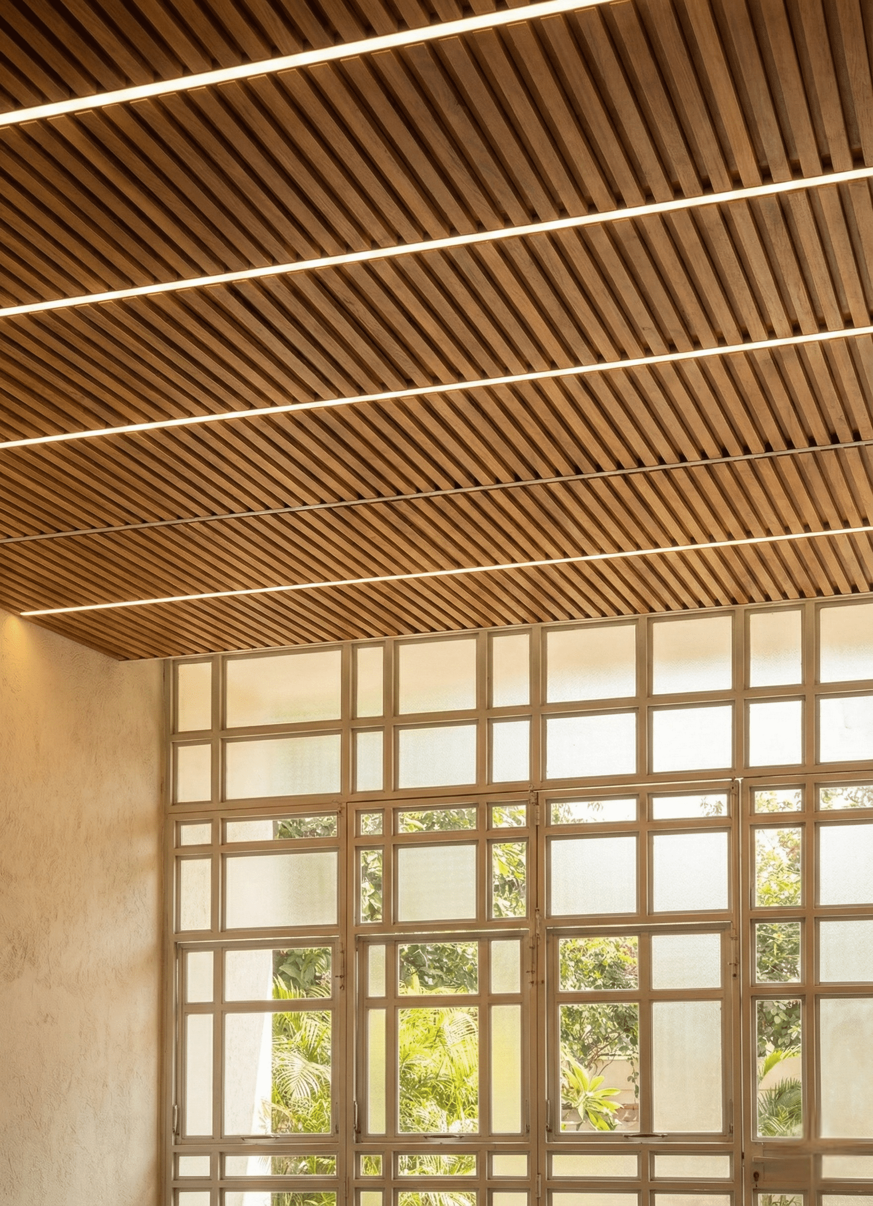 Thiết kế trần nhà ốp thanh lam gỗ song song kết hợp các dải đèn LED âm trần chạy ngang tạo điểm nhấn hiện đại, ấm cúng. Bên dưới là hệ vách kính khung chia ô lớn giúp lấy sáng tự nhiên tối đa và mở rộng tầm nhìn ra sân vườn xanh mát